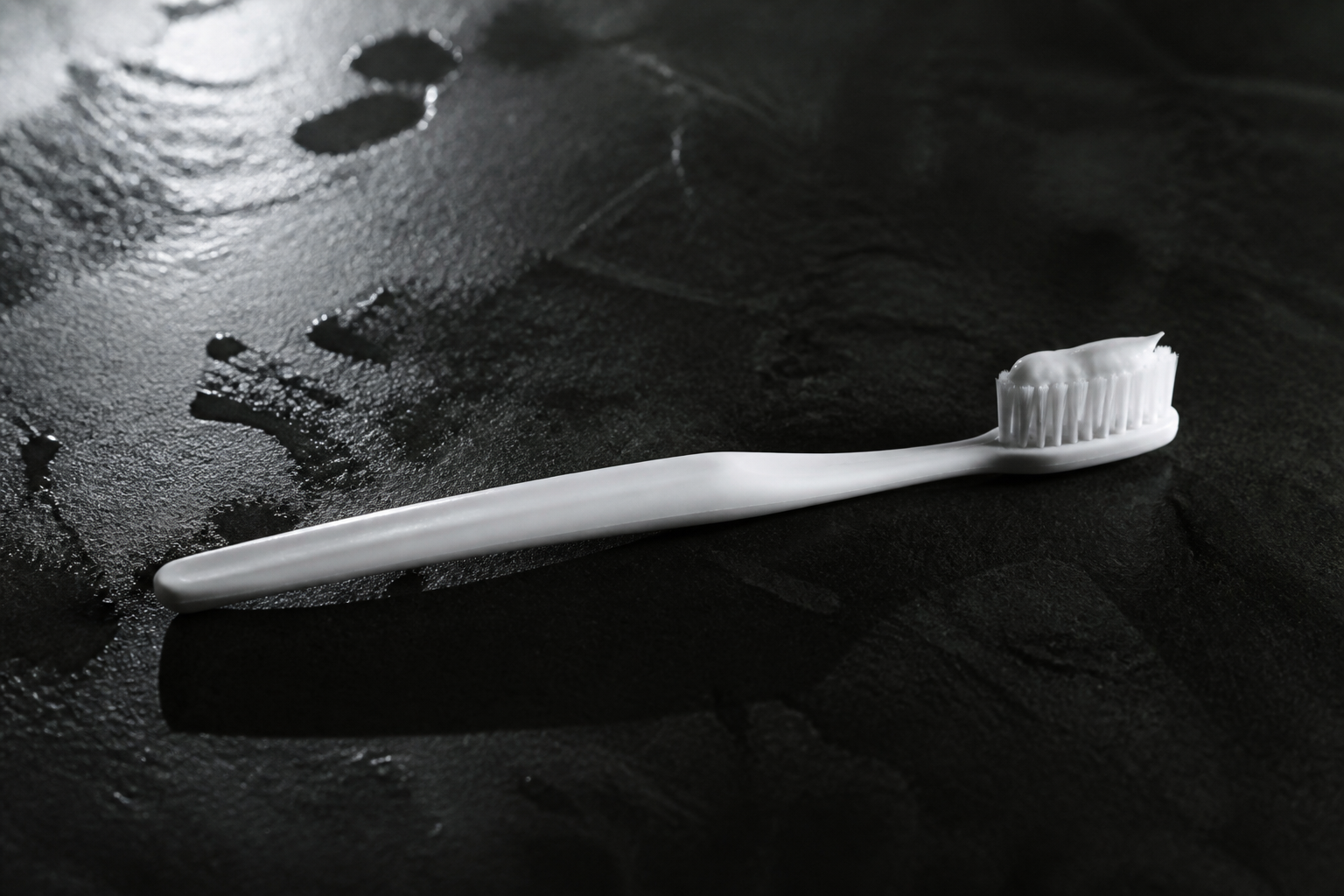 A white toothbrush with paste on a dark wet bathroom counter